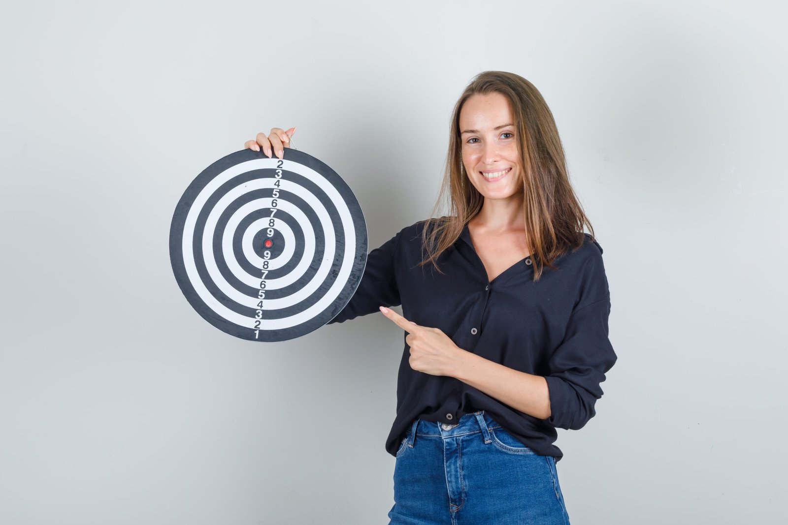 Young woman pointing finger at dartboard in black shirt, jeans shorts and looking cheerful , front view.